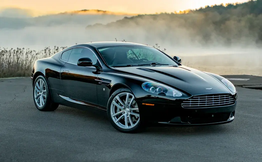 A sleek black sports car is parked on a misty road at sunrise. The car's shiny exterior and sharp design elements stand out against the serene landscape.