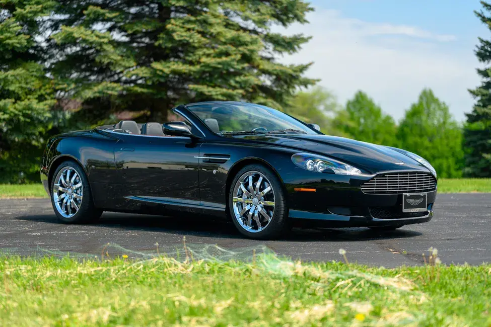 A sleek black convertible sports car with shiny chrome wheels parked on pavement, set against a lush green park landscape with trees under a clear sky.
