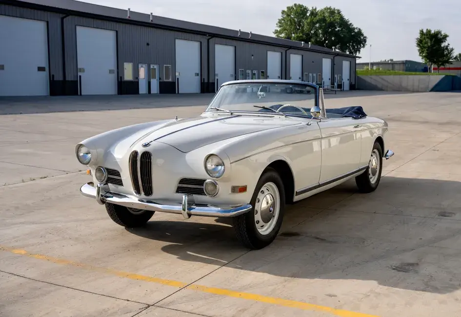 A classic white convertible car parked on a concrete lot in front of a row of industrial garage doors, with trees and grass in the background.