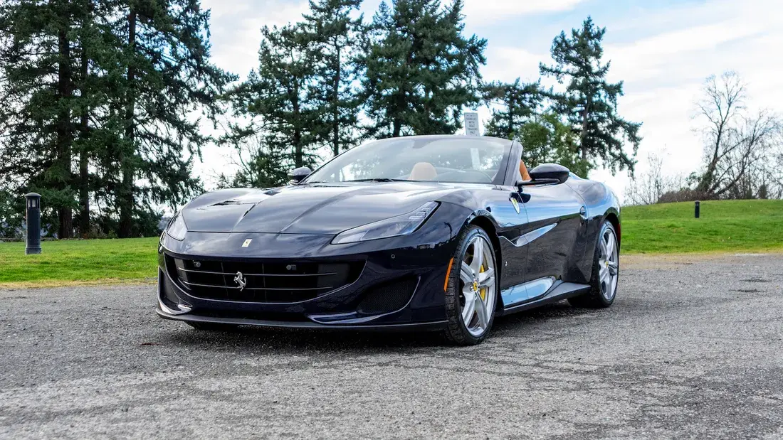 A sleek black convertible sports car with silver rims is parked on a paved surface. The background features lush green grass and tall trees under a partly cloudy sky.