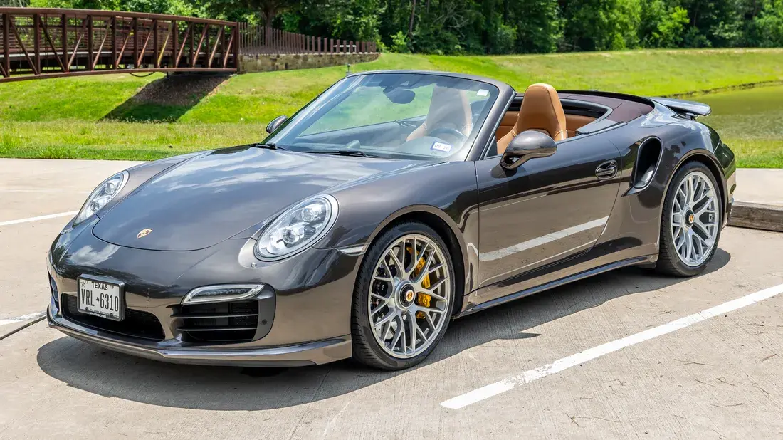 A dark gray Porsche 911 convertible with tan leather seats and silver multi-spoke wheels parked on a concrete lot near a grassy area and a bridge.