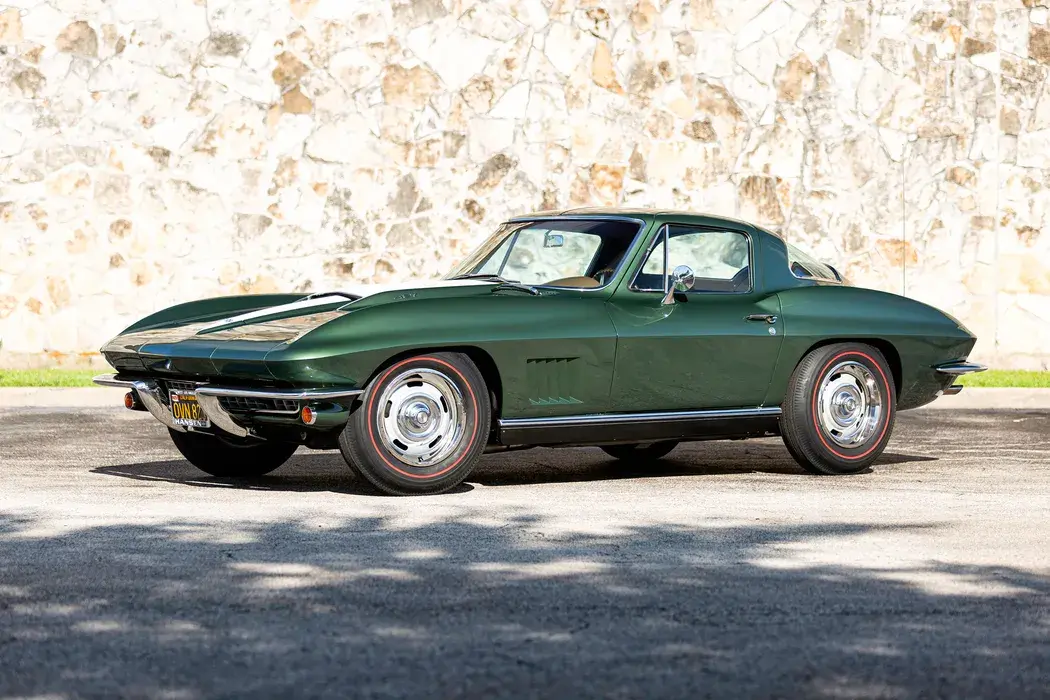 A classic green sports car with chrome accents and round headlights is parked on a street. A textured stone wall serves as the background.